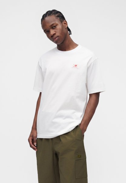 Joven con cabello trenzado que lleva una camiseta blanca de New Balance y pantalones cargo verde oliva, de pie con una mano en el bolsillo.