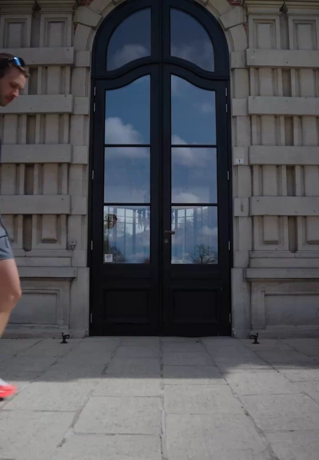 Hombre con gafas de sol y pantalones cortos caminando junto a una puerta de cristal alta con marco negro en la fachada de un edificio de piedra gris.