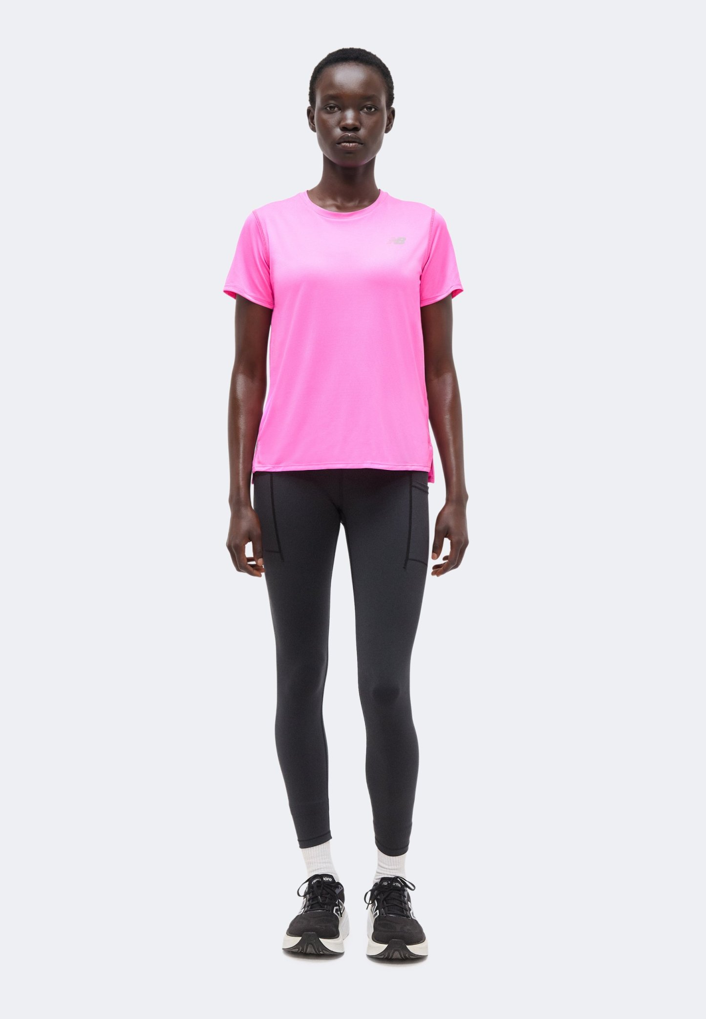 Mujer de pie erguida vistiendo una camiseta de manga corta rosa brillante, leggings negros, calcetines blancos y zapatillas deportivas negras sobre un fondo liso.