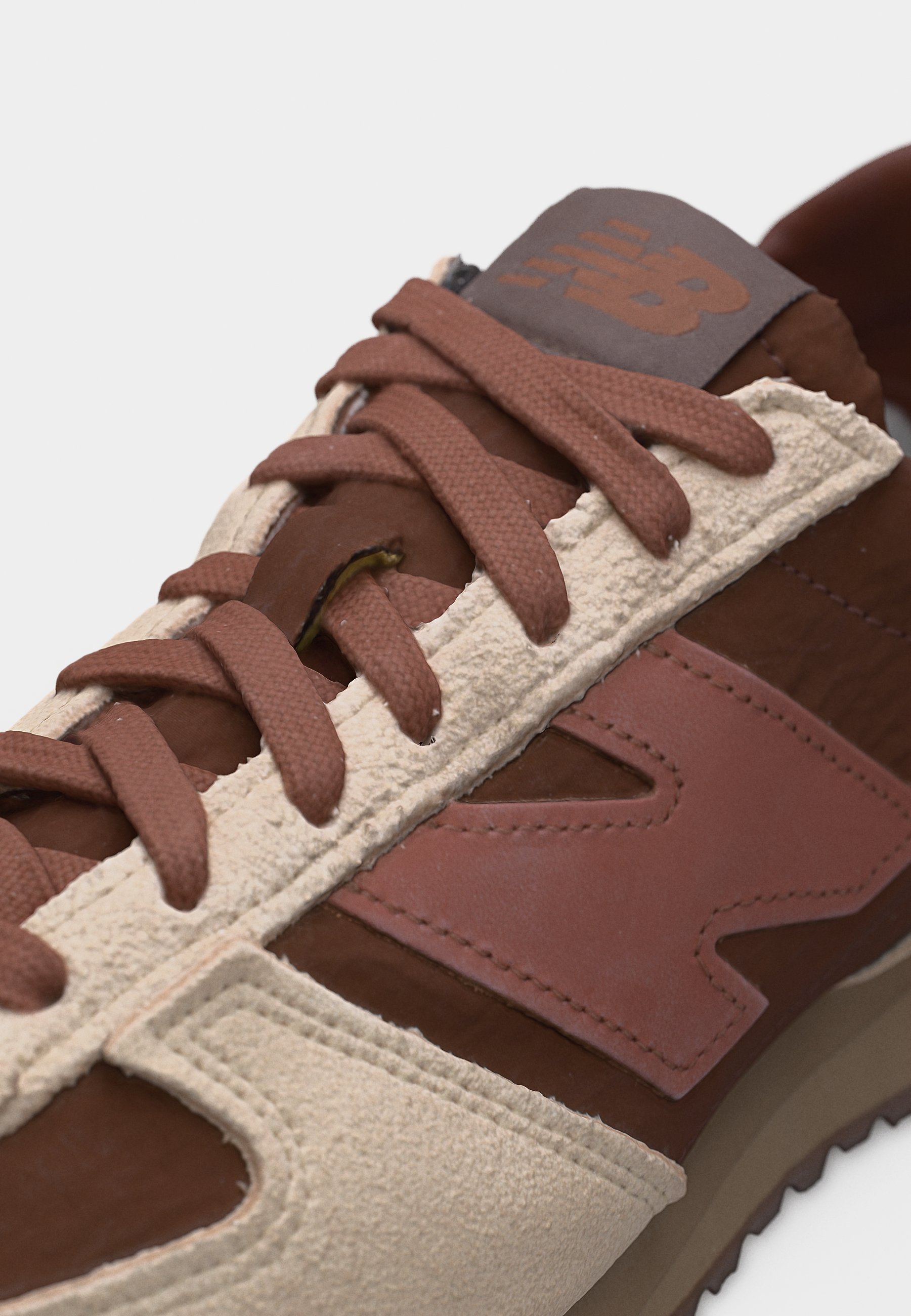 Zapatilla New Balance en tonos marrón y beige con paneles de ante y cuero, cordones marrones y un logotipo visible cosido en el lateral y la lengüeta.