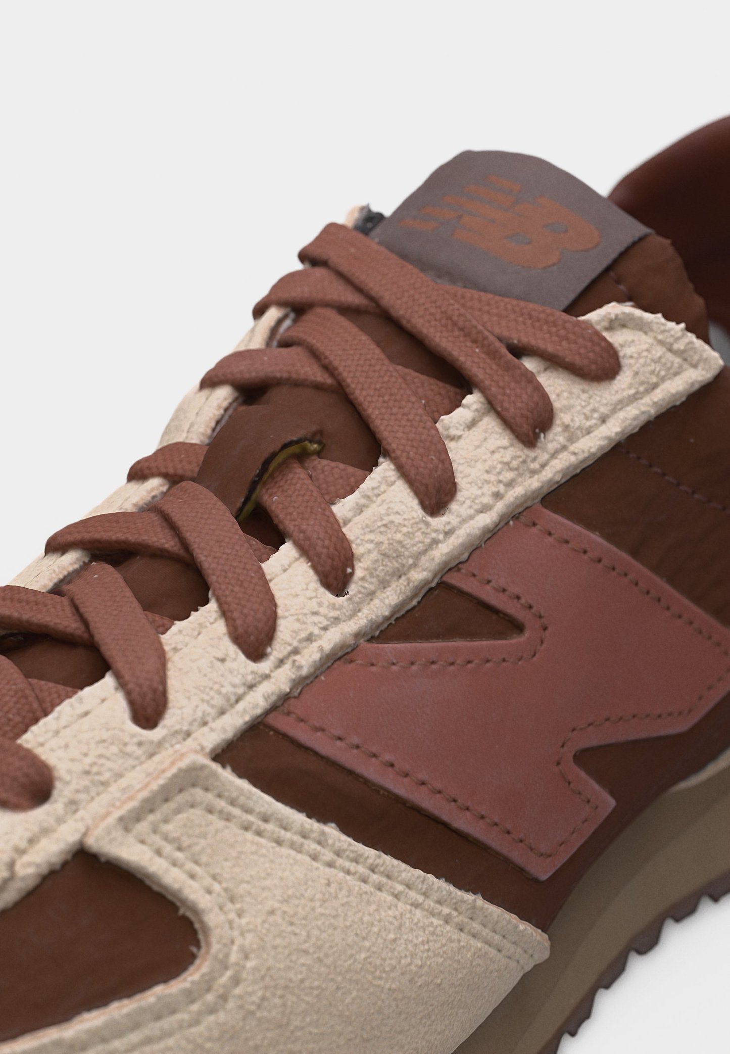 Zapatilla New Balance en tonos marrón y beige con paneles de ante y cuero, cordones marrones y un logotipo visible cosido en el lateral y la lengüeta.