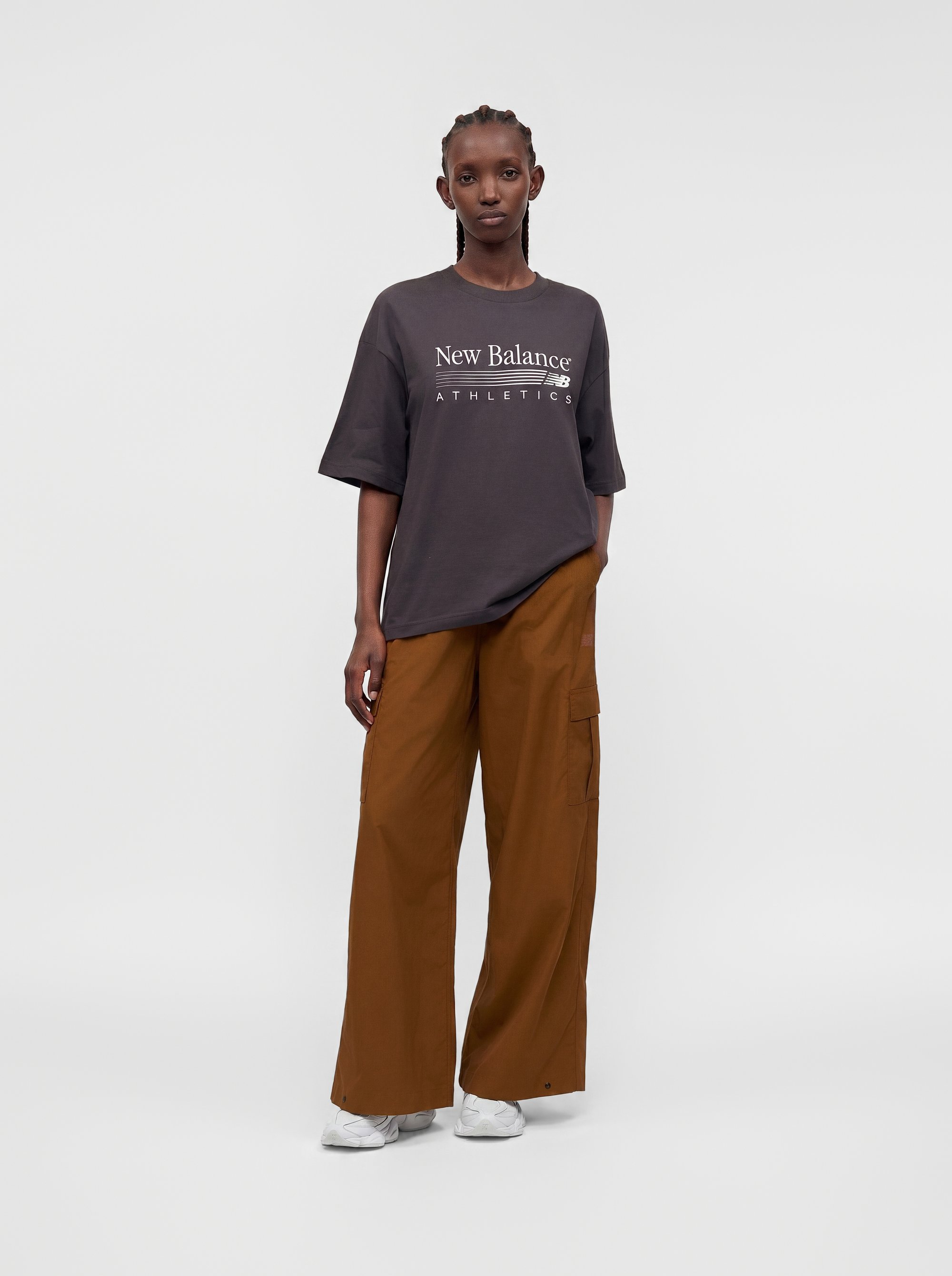 Persona con una camiseta oscura y oversize de New Balance Athletics, pantalones marrones de pierna ancha y zapatillas blancas, de pie frente a un fondo claro y liso.
