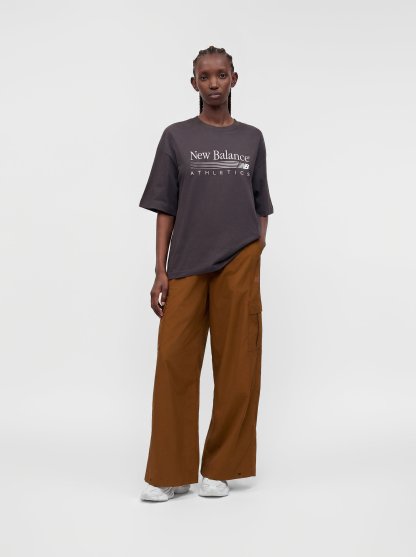 Persona con una camiseta oscura y oversize de New Balance Athletics, pantalones marrones de pierna ancha y zapatillas blancas, de pie frente a un fondo claro y liso.