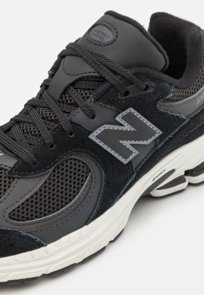 Zapatilla deportiva negra con paneles de malla y ante, con un destacado logotipo plateado "N", cordones texturizados y una suela de caucho blanca.