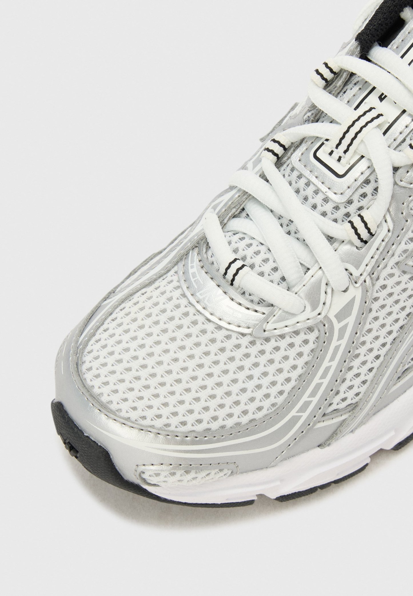 Primer plano de una zapatilla deportiva blanca y plateada con parte superior de malla, cordones blancos con rayas negras y suela de goma en blanco y negro.