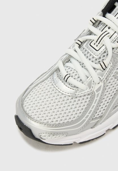 Primer plano de una zapatilla deportiva blanca y plateada con parte superior de malla, cordones blancos con rayas negras y suela de goma en blanco y negro.