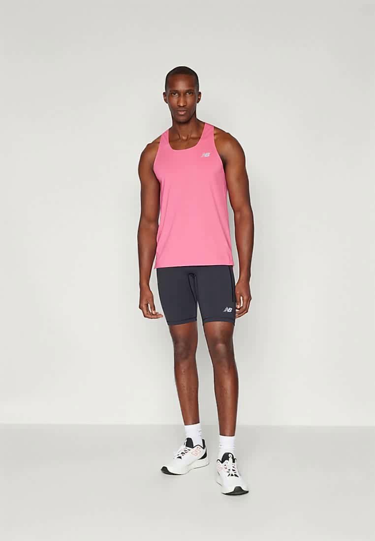 Hombre de pie con camiseta de atletismo sin mangas de color rosa, pantalones cortos ajustados negros, calcetines blancos y zapatillas de correr blancas, contra un fondo claro y simple.