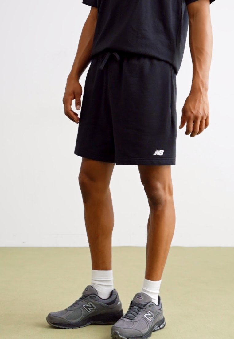 Pantalones cortos deportivos negros con cintura elástica y cordón. Combinados con zapatillas New Balance grises y calcetines blancos de caña media. Diseño sencillo.