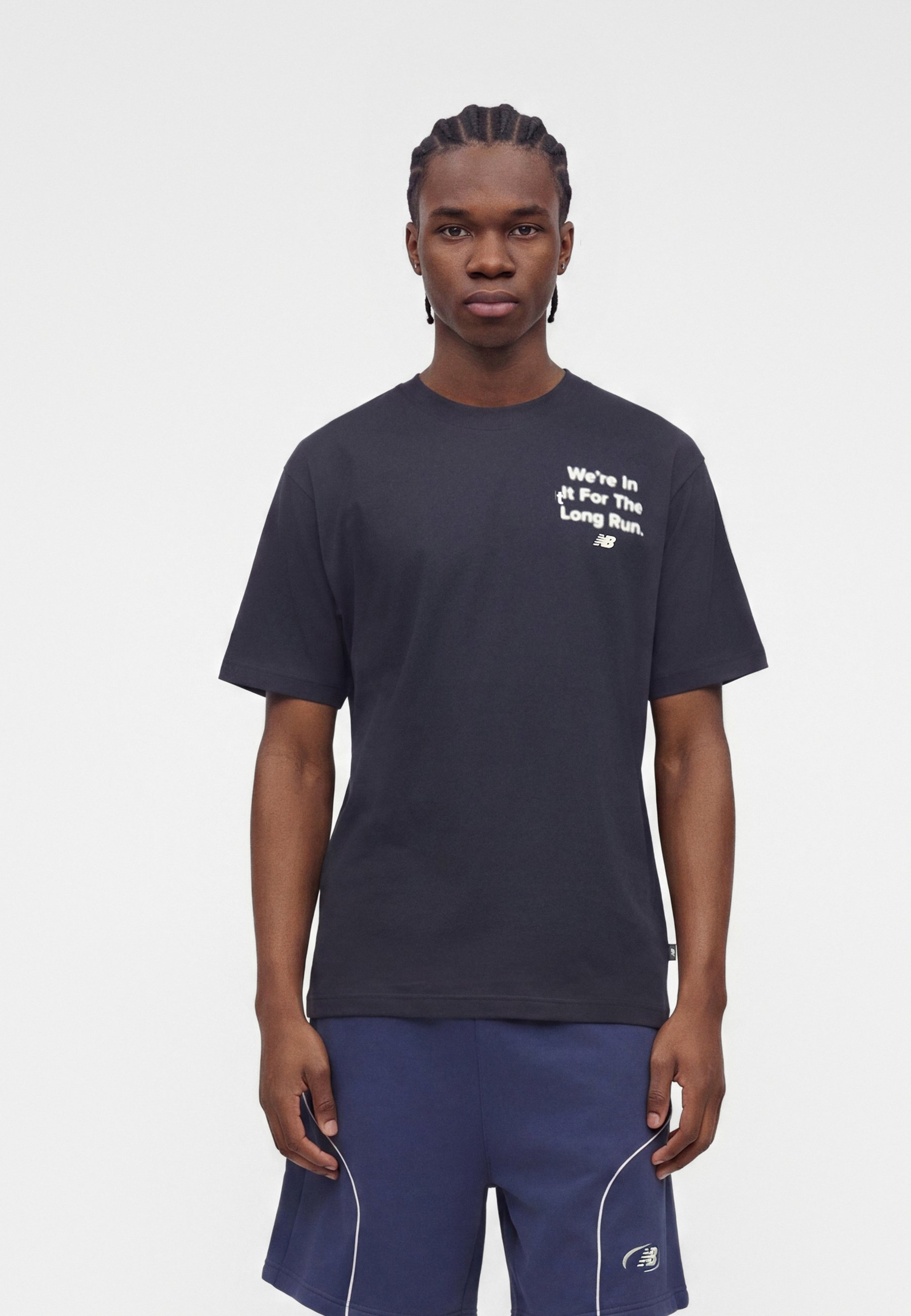 Joven con el pelo trenzado que lleva una camiseta negra con texto blanco y pantalones cortos deportivos azul marino sobre un fondo blanco liso.