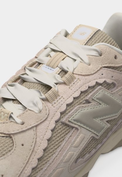 Primer plano de zapatilla New Balance beige mostrando texturas de malla y ante, cordones anchos de satén, y el logotipo reflectante "N" de la marca en el lateral.