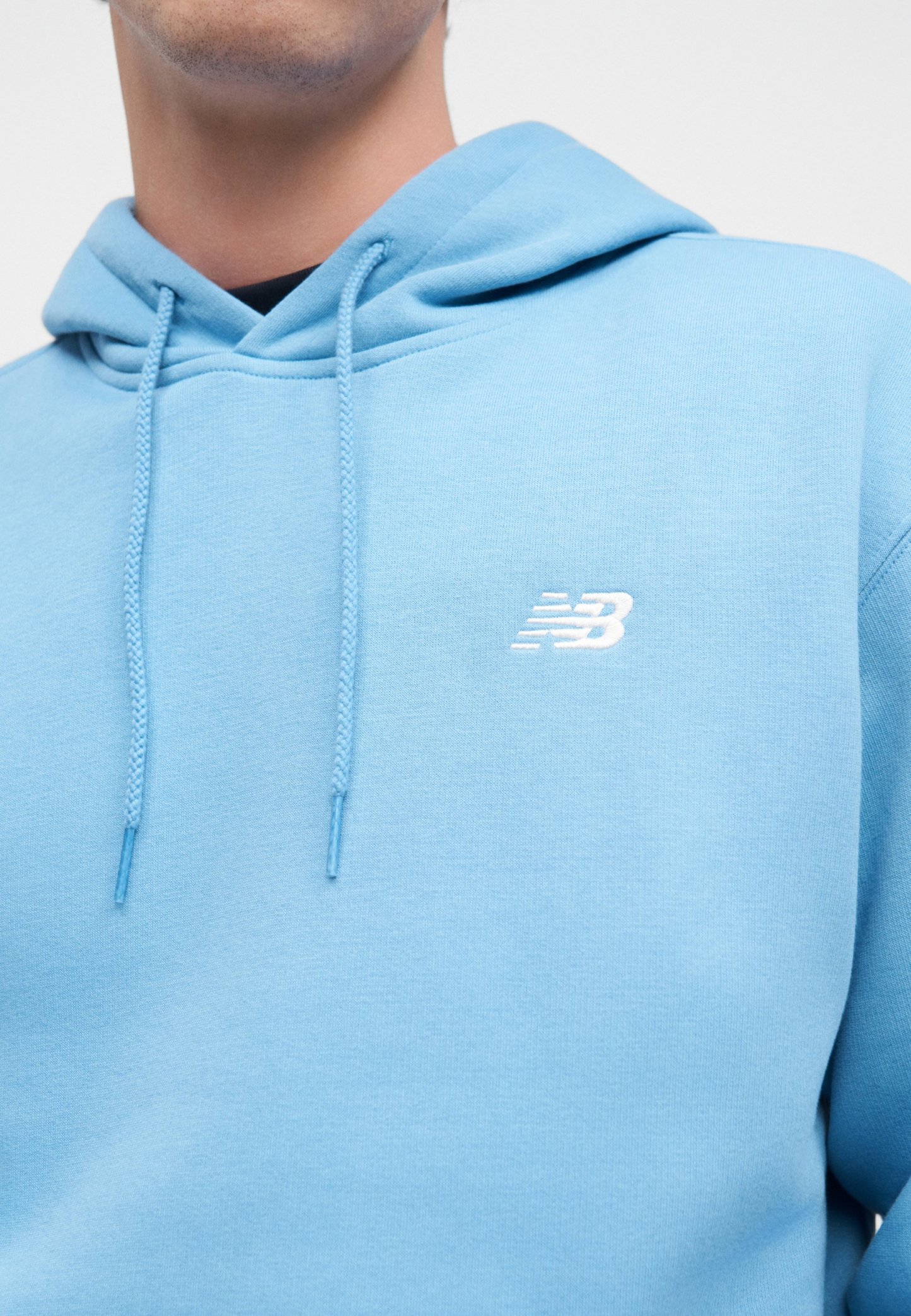 Hombre con sudadera con capucha azul claro con cordones y un pequeño logo blanco de New Balance en el pecho sobre un fondo sencillo.