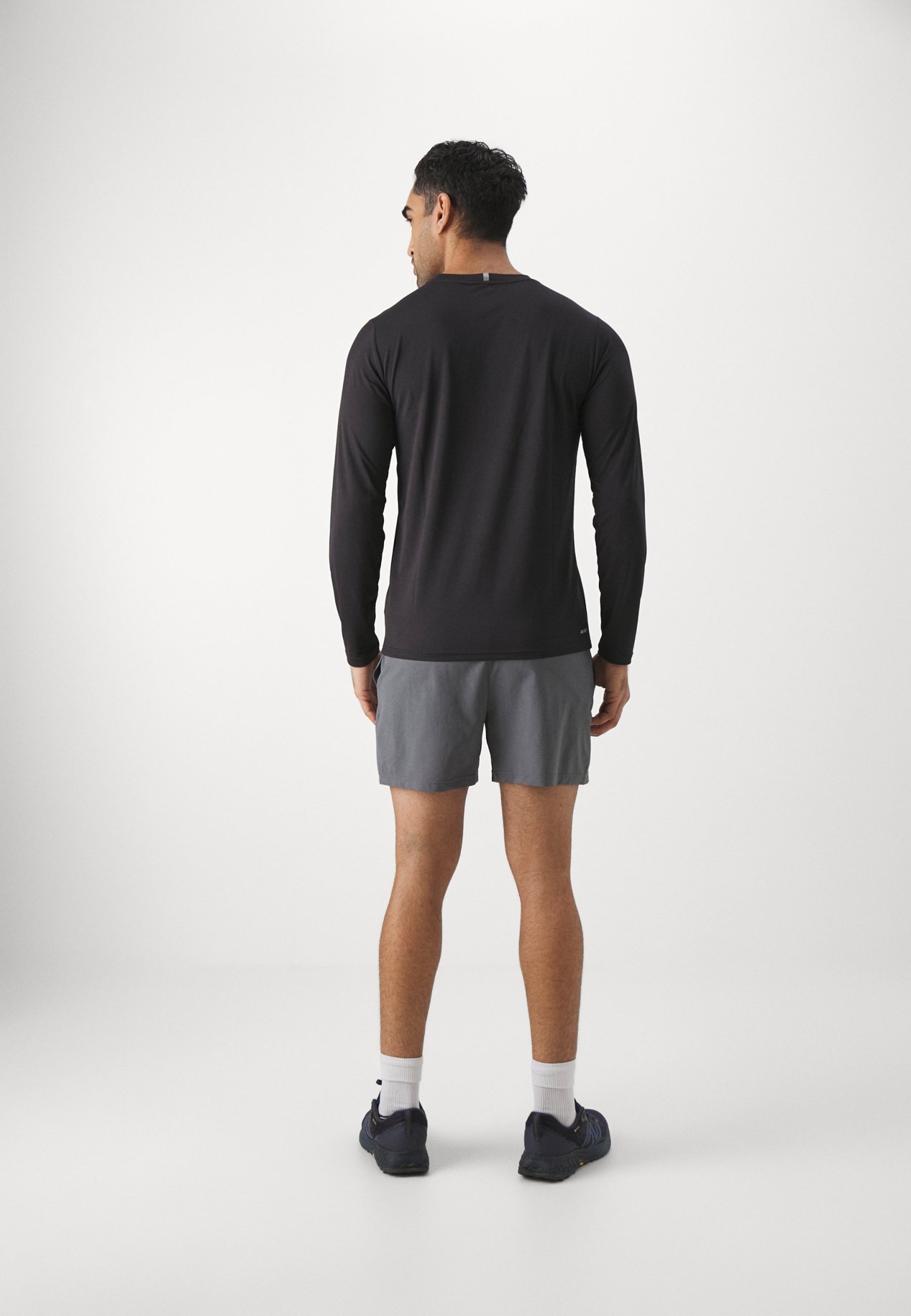 Camiseta de manga larga negra para hombre con cuello redondo, combinada con pantalones cortos grises. La persona se presenta de espaldas, mostrando un diseño ajustado y calzado deportivo.