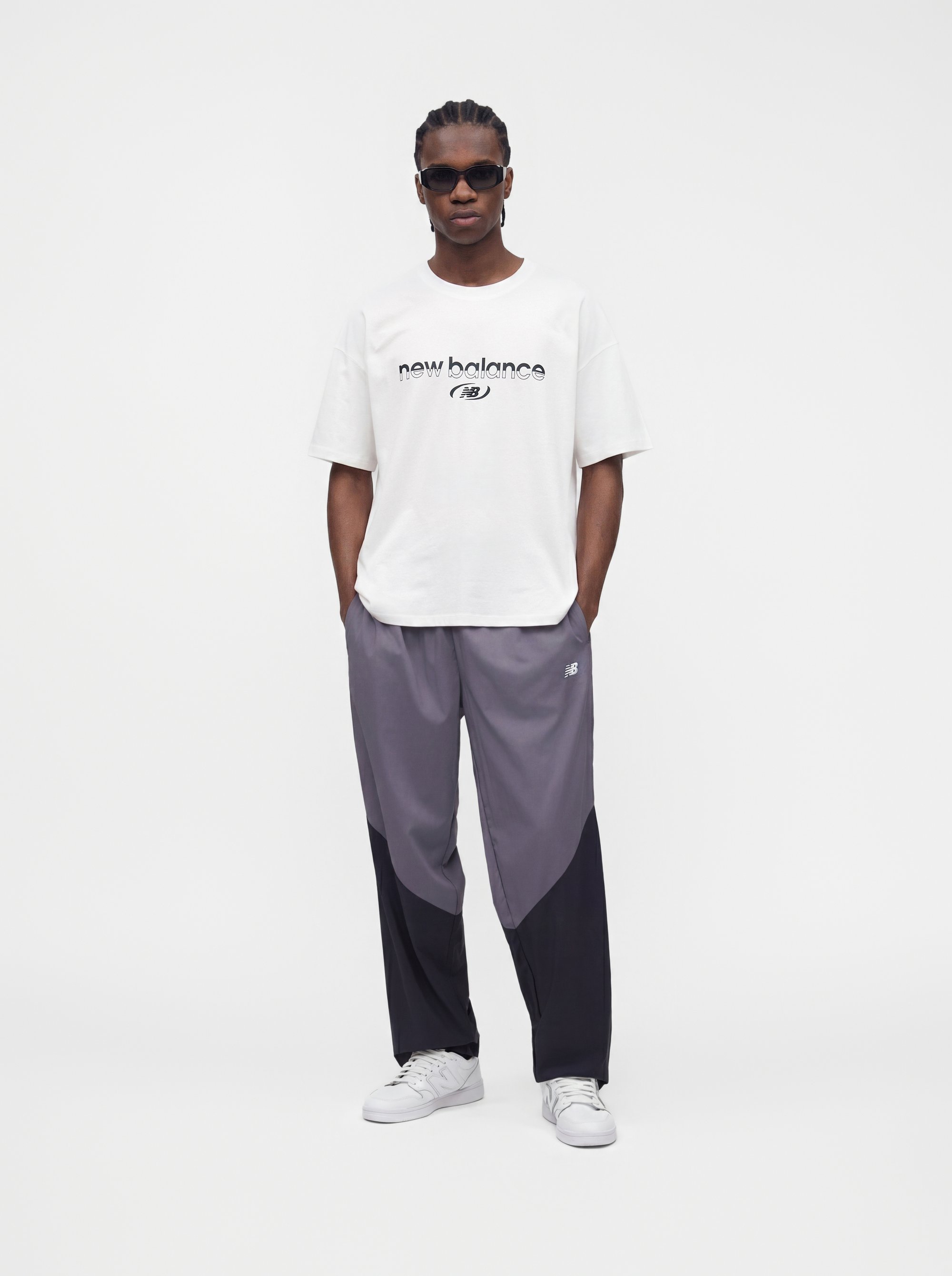 Hombre con cabello trenzado y gafas de sol, vistiendo una camiseta blanca de New Balance, pantalones deportivos gris bicolor y zapatillas blancas, de pie con las manos en los bolsillos.
