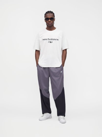 Hombre con cabello trenzado y gafas de sol, vistiendo una camiseta blanca de New Balance, pantalones deportivos gris bicolor y zapatillas blancas, de pie con las manos en los bolsillos.