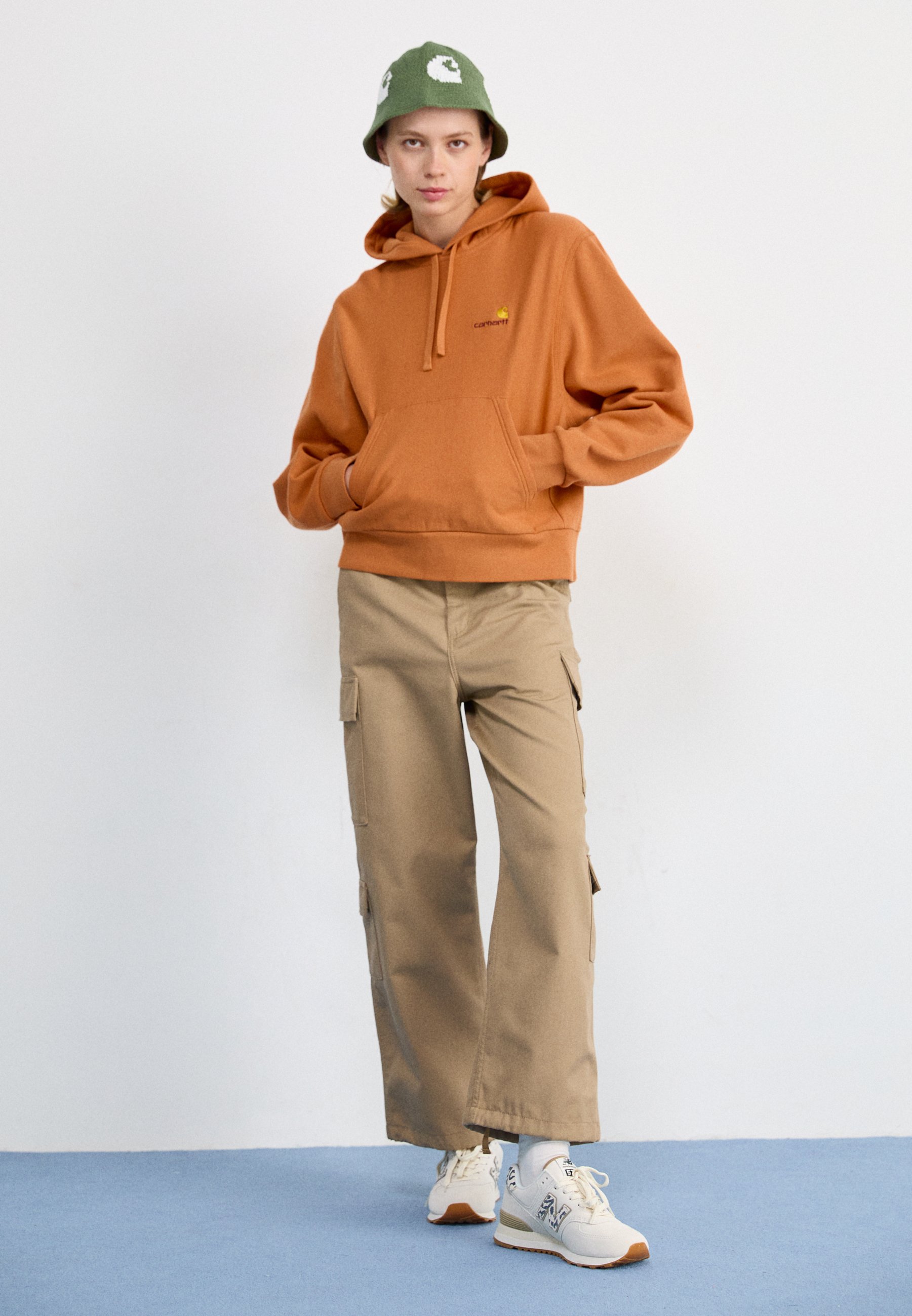 Persona joven con sudadera con capucha naranja, pantalones cargo beige, zapatillas blancas y un sombrero tipo cubo verde, de pie contra un fondo claro y uniforme.