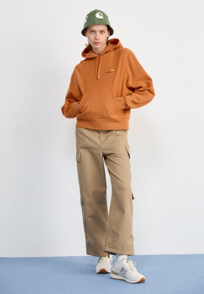 Persona joven con sudadera con capucha naranja, pantalones cargo beige, zapatillas blancas y un sombrero tipo cubo verde, de pie contra un fondo claro y uniforme.