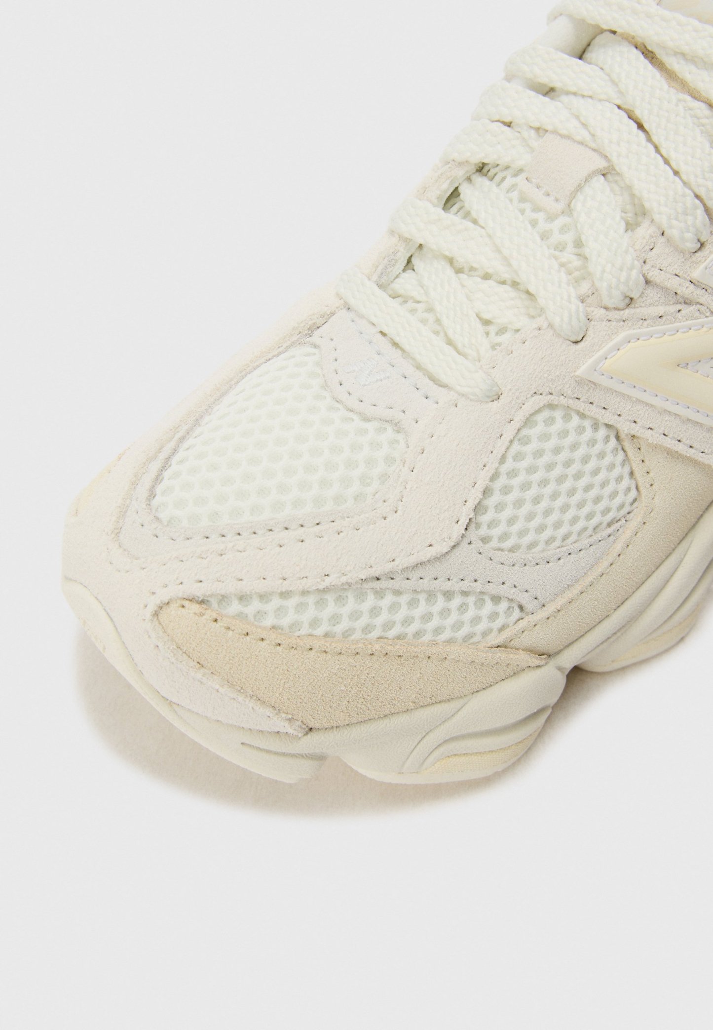 Primer plano de la punta de zapatilla de malla blanca y beige con superposiciones de ante y cordones gruesos fuertemente atados sobre fondo claro.