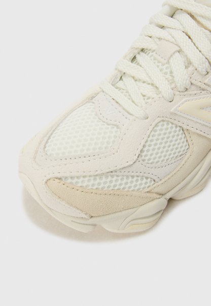 Primer plano de la punta de zapatilla de malla blanca y beige con superposiciones de ante y cordones gruesos fuertemente atados sobre fondo claro.