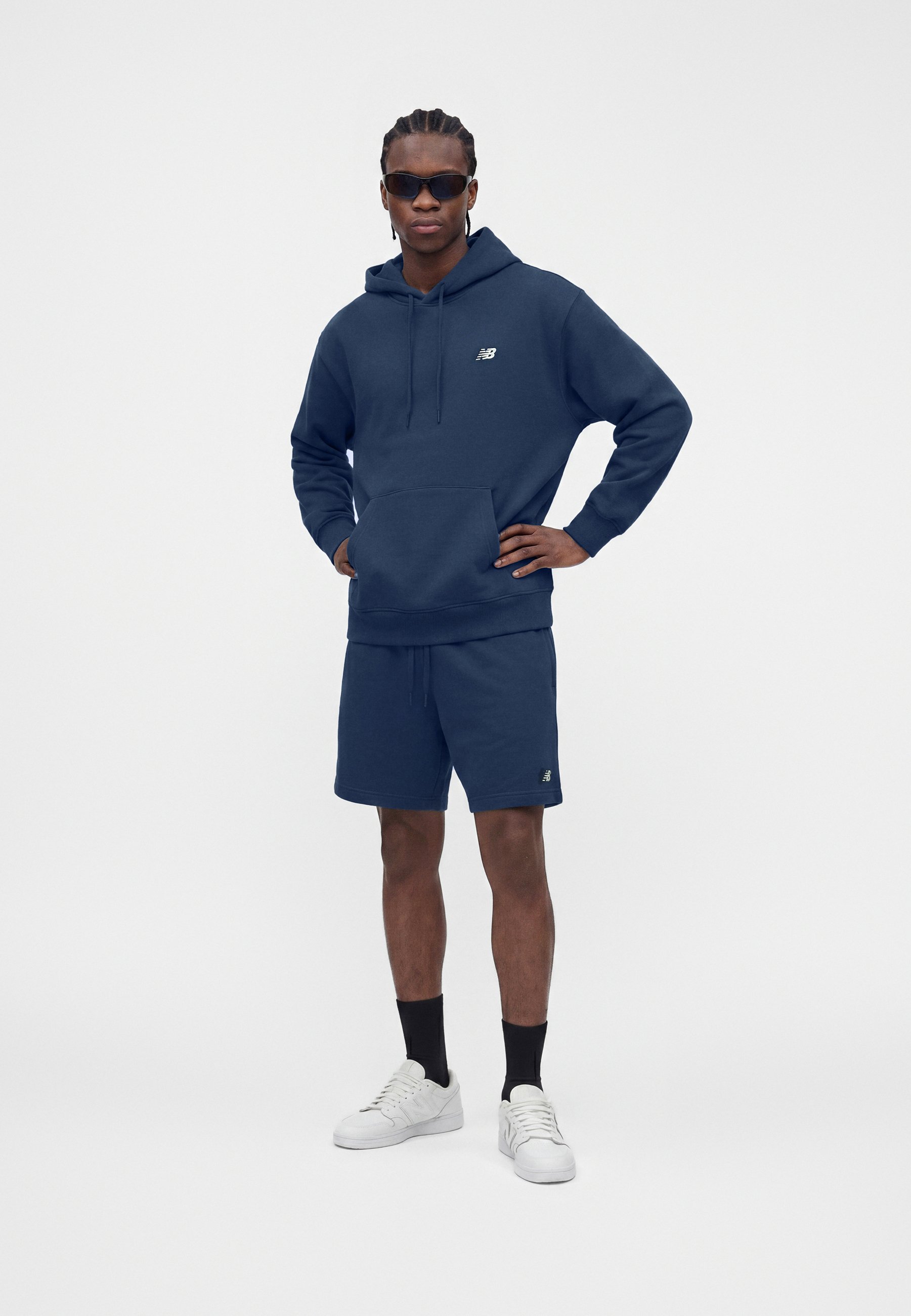Hombre con sudadera con capucha azul marino y pantalones cortos, zapatillas blancas, calcetines negros y gafas de sol oscuras, de pie con una mano en la cadera.