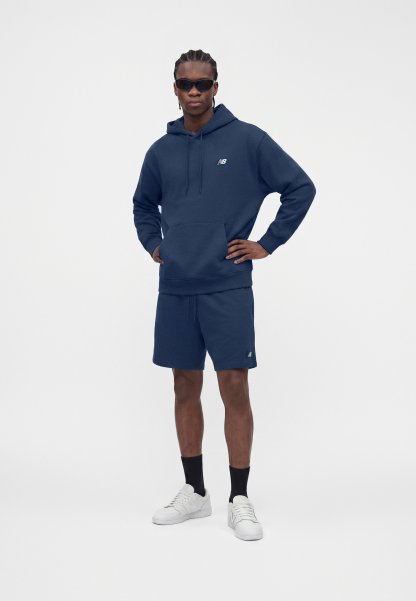 Hombre con sudadera con capucha azul marino y pantalones cortos, zapatillas blancas, calcetines negros y gafas de sol oscuras, de pie con una mano en la cadera.