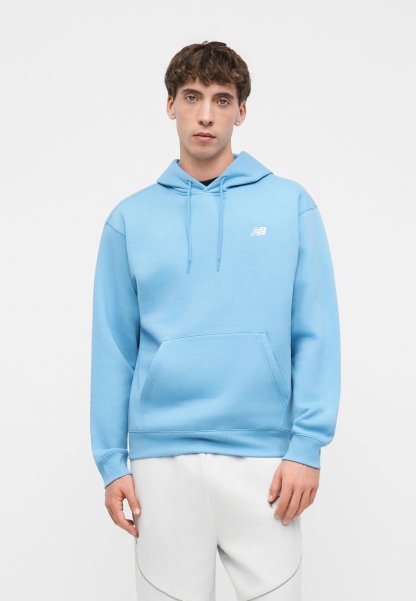 Joven con sudadera con capucha azul clara con bolsillo canguro y pantalones blancos, de pie sobre un fondo blanco liso.