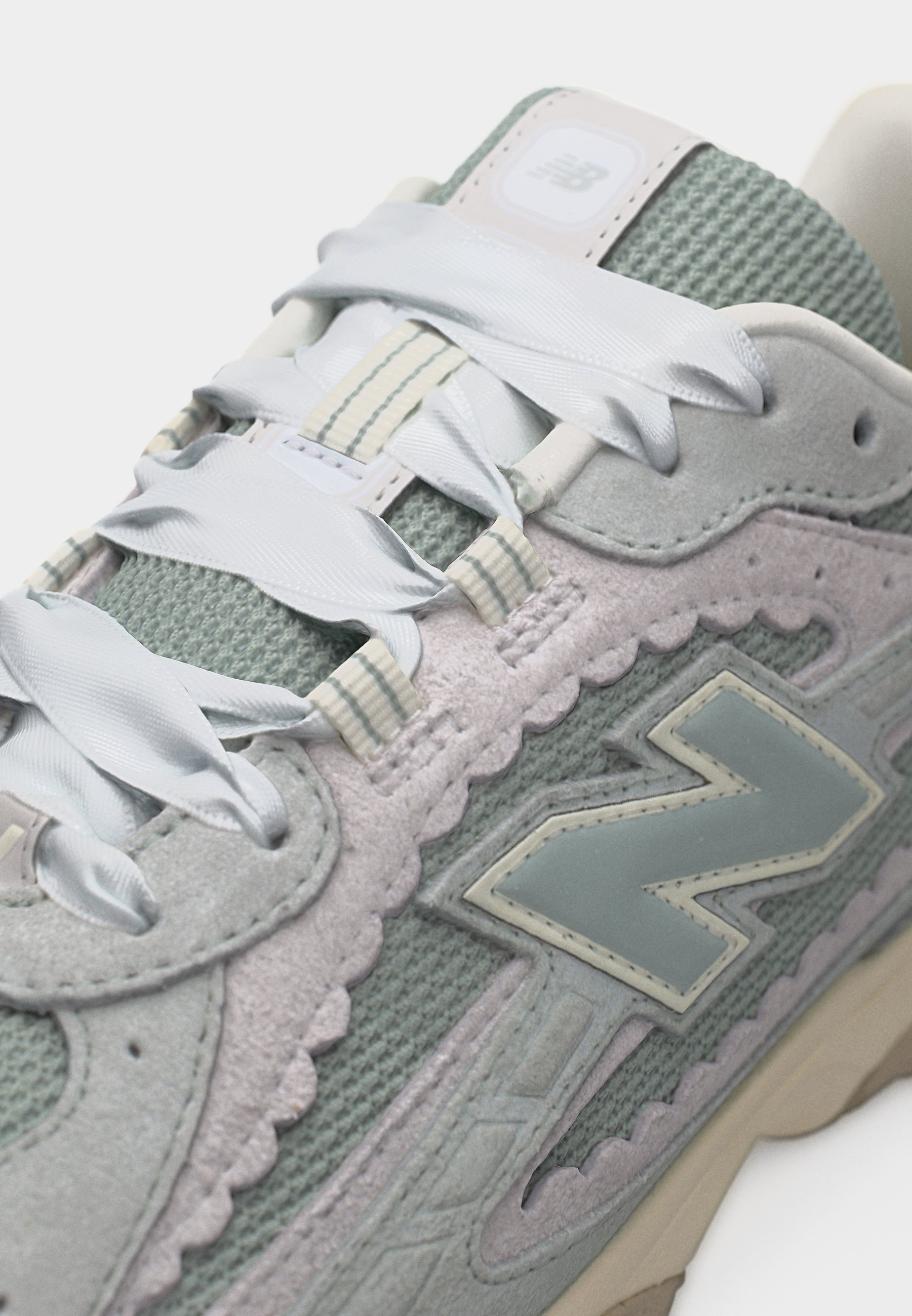 Primer plano de una zapatilla New Balance gris y verde claro con cordones blancos anchos, parte superior de malla, superposiciones de ante y un gran logo "N" en el lateral.