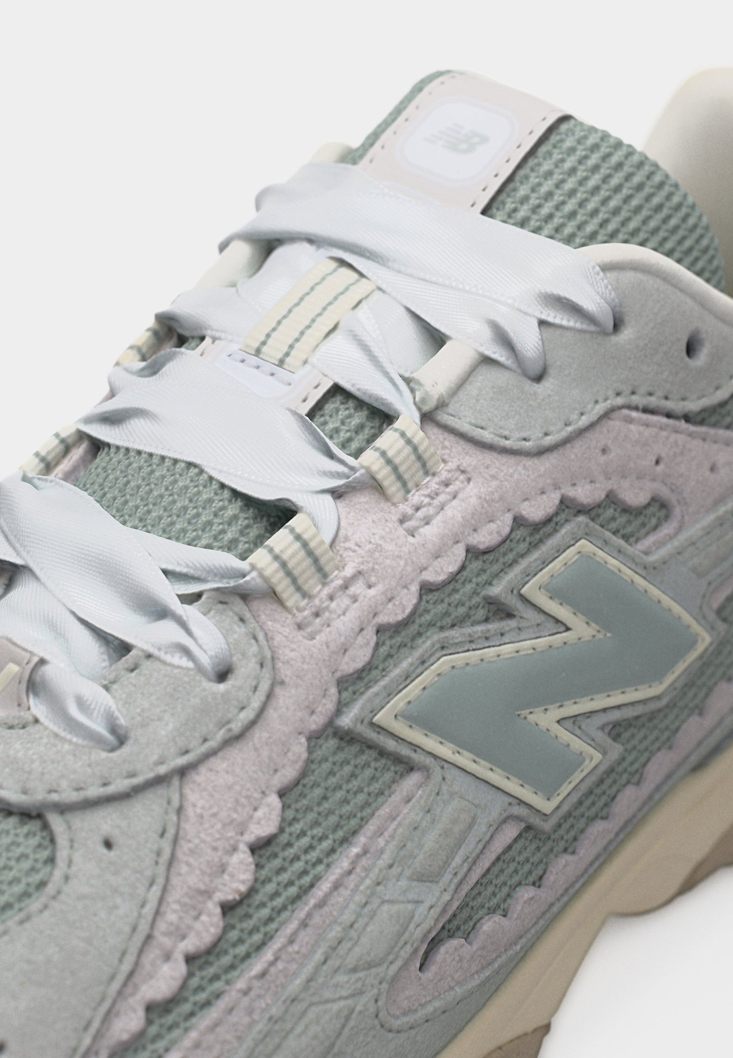 Primer plano de una zapatilla New Balance gris y verde claro con cordones blancos anchos, parte superior de malla, superposiciones de ante y un gran logo "N" en el lateral.