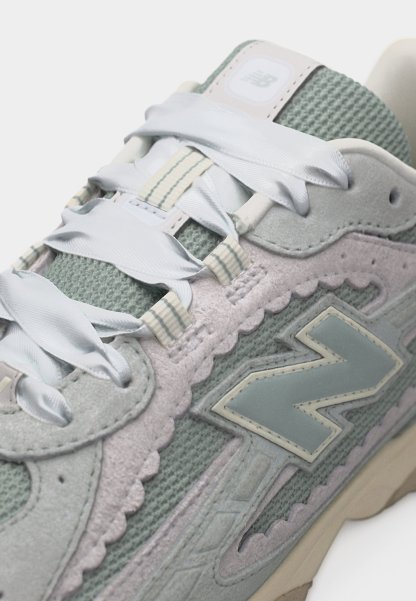 Primer plano de una zapatilla New Balance gris y verde claro con cordones blancos anchos, parte superior de malla, superposiciones de ante y un gran logo "N" en el lateral.