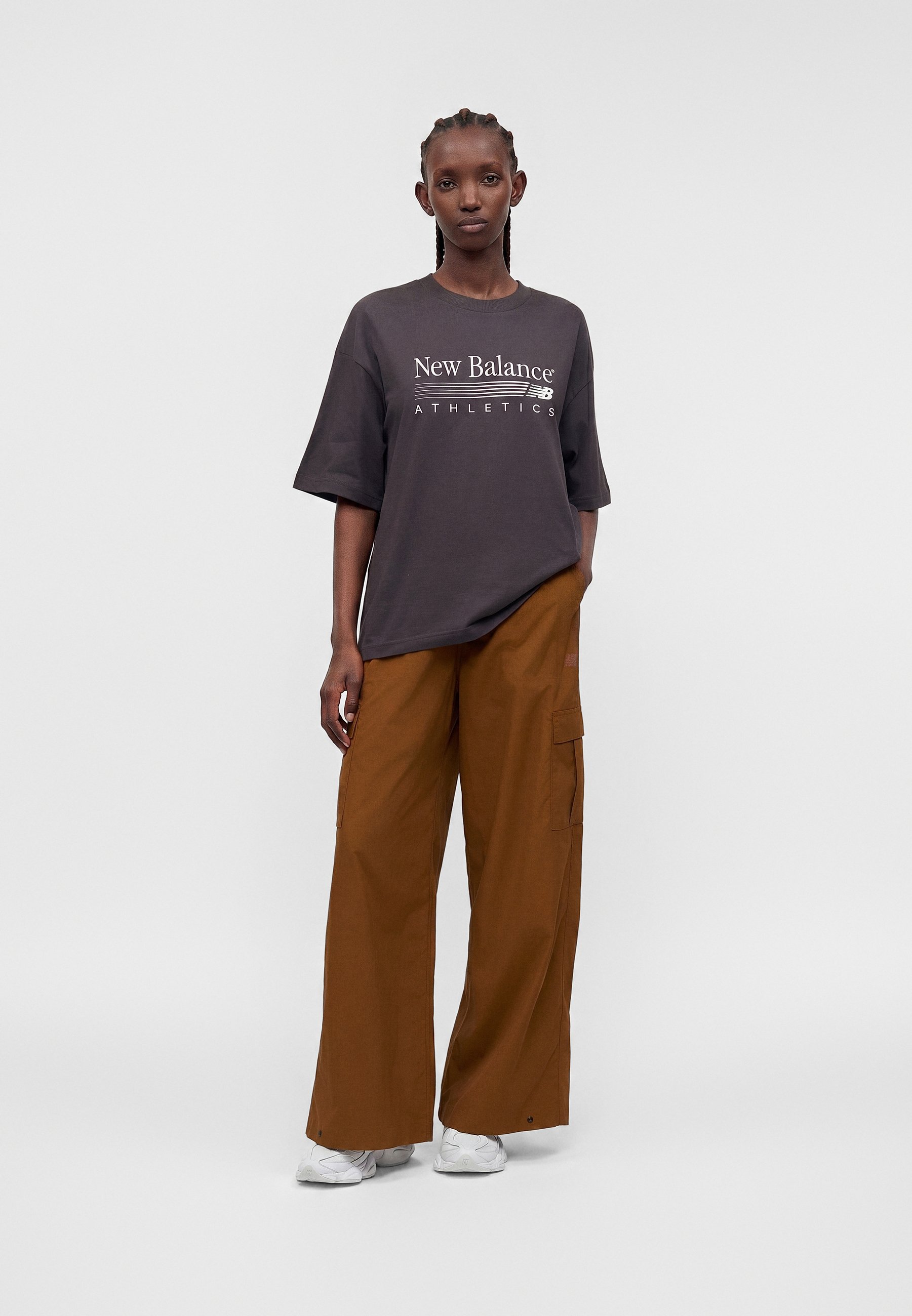 Mujer con trenzas, vestida con una camiseta de New Balance Athletics gris oscuro, pantalones anchos marrones y zapatillas blancas, de pie frente a un fondo gris.