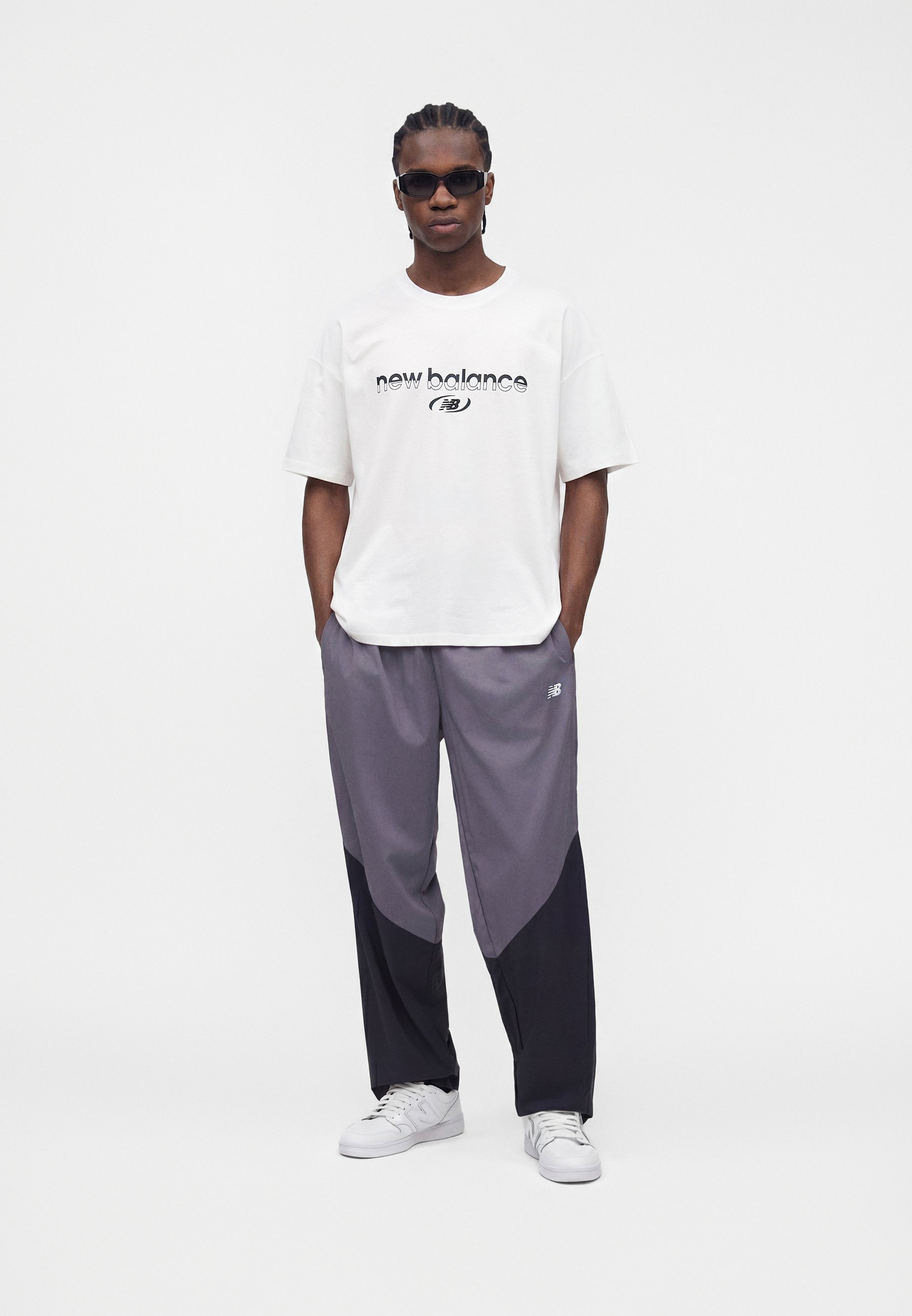 Hombre con gafas de sol negras, camiseta blanca de New Balance, pantalones de chándal bicolor gris y negro, y zapatillas blancas, de pie con las manos en los bolsillos.