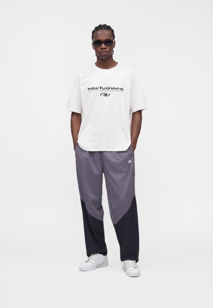 Hombre con gafas de sol negras, camiseta blanca de New Balance, pantalones de chándal bicolor gris y negro, y zapatillas blancas, de pie con las manos en los bolsillos.