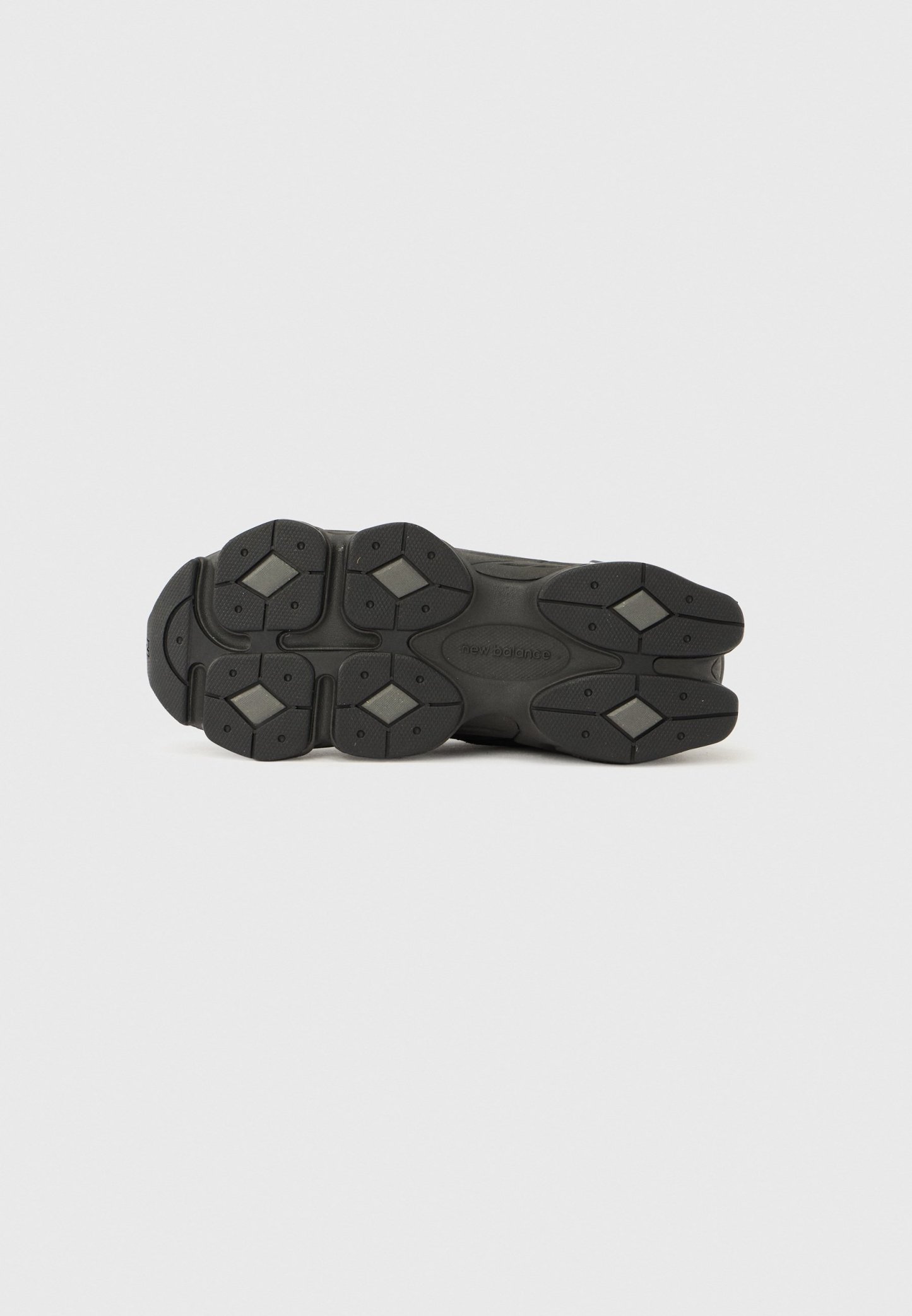 Suela de zapato deportivo negra con superficie texturizada, múltiples formas contorneadas y patrones de agarre de goma hexagonales. Etiquetada "new balance."