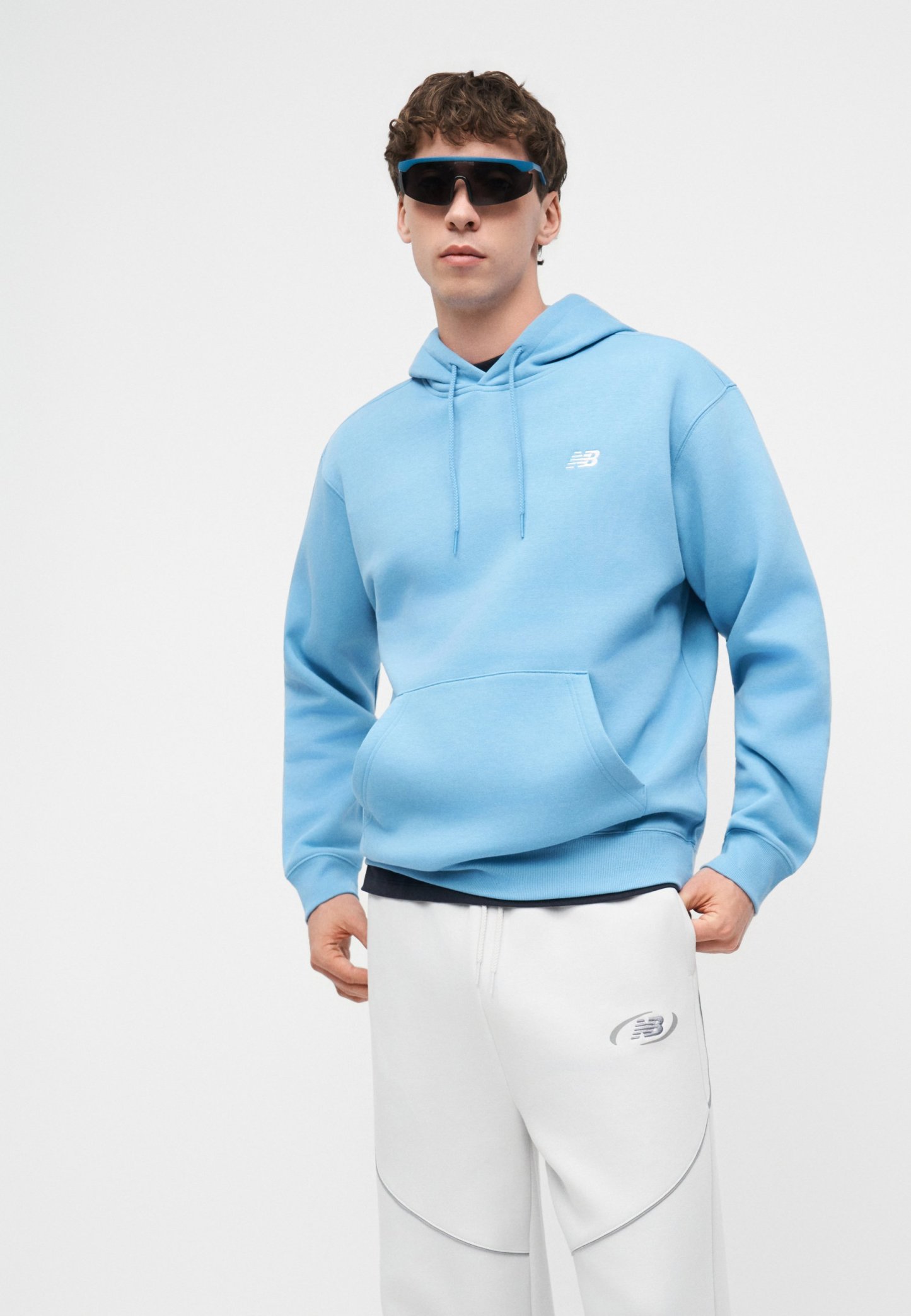 Joven con sudadera azul con capucha, pantalones blancos y grandes gafas de sol deportivas negras sobre un fondo blanco liso.