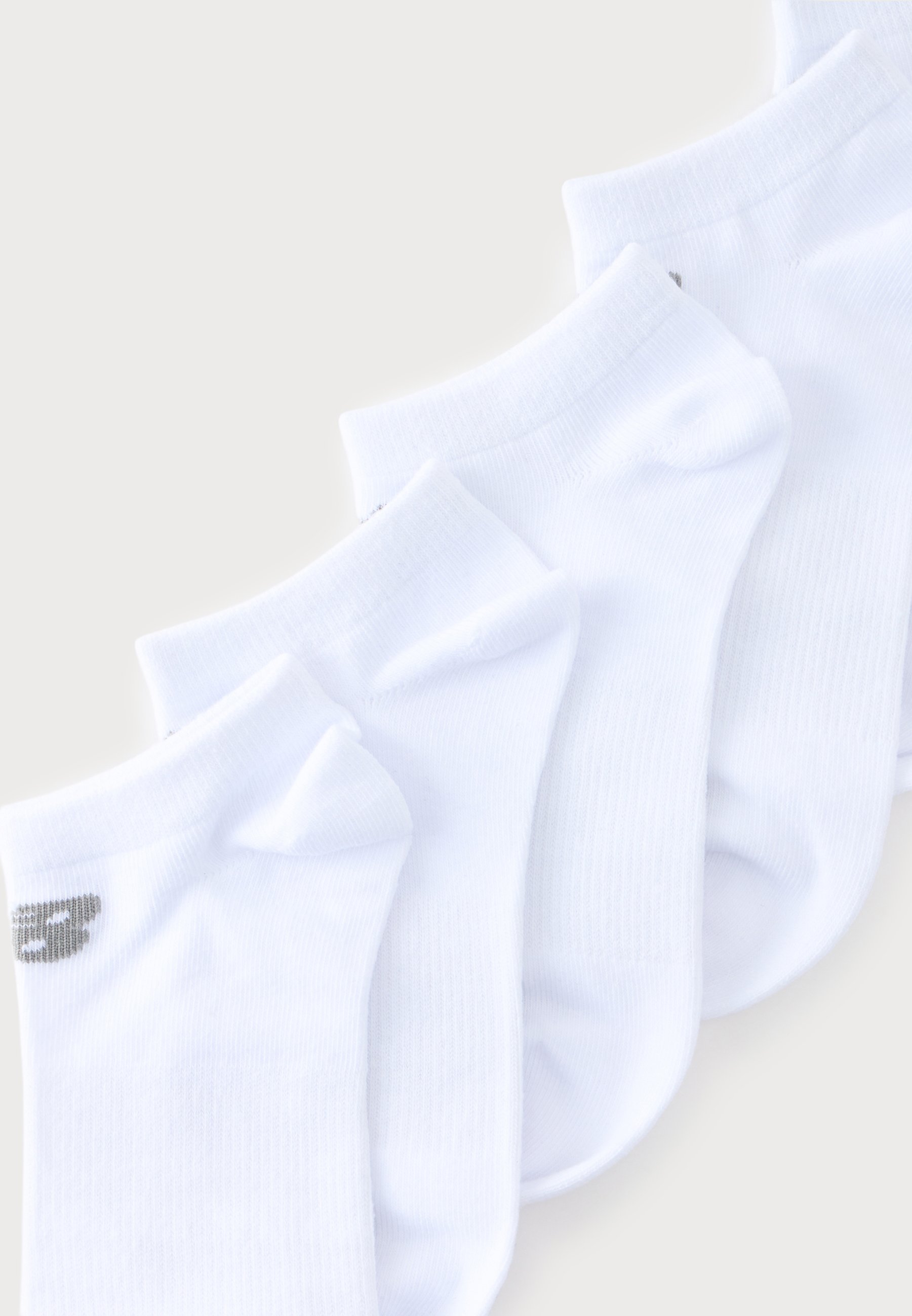 Cuatro pares de calcetines blancos tobilleros dispuestos en diagonal, con un pequeño logo gris visible en la puntera del par más delantero.