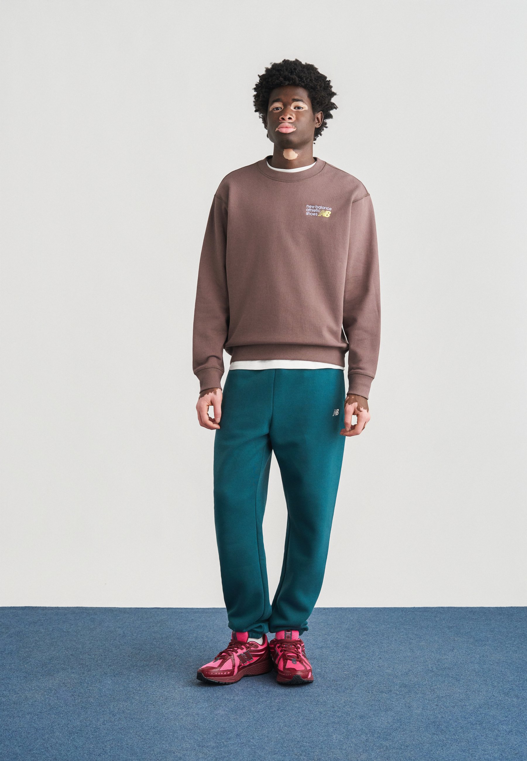 Sudadera marrón con logo bordado, pantalones deportivos verde azulado y zapatillas deportivas rosas. La tela es suave; el diseño es de corte relajado con puños acanalados.