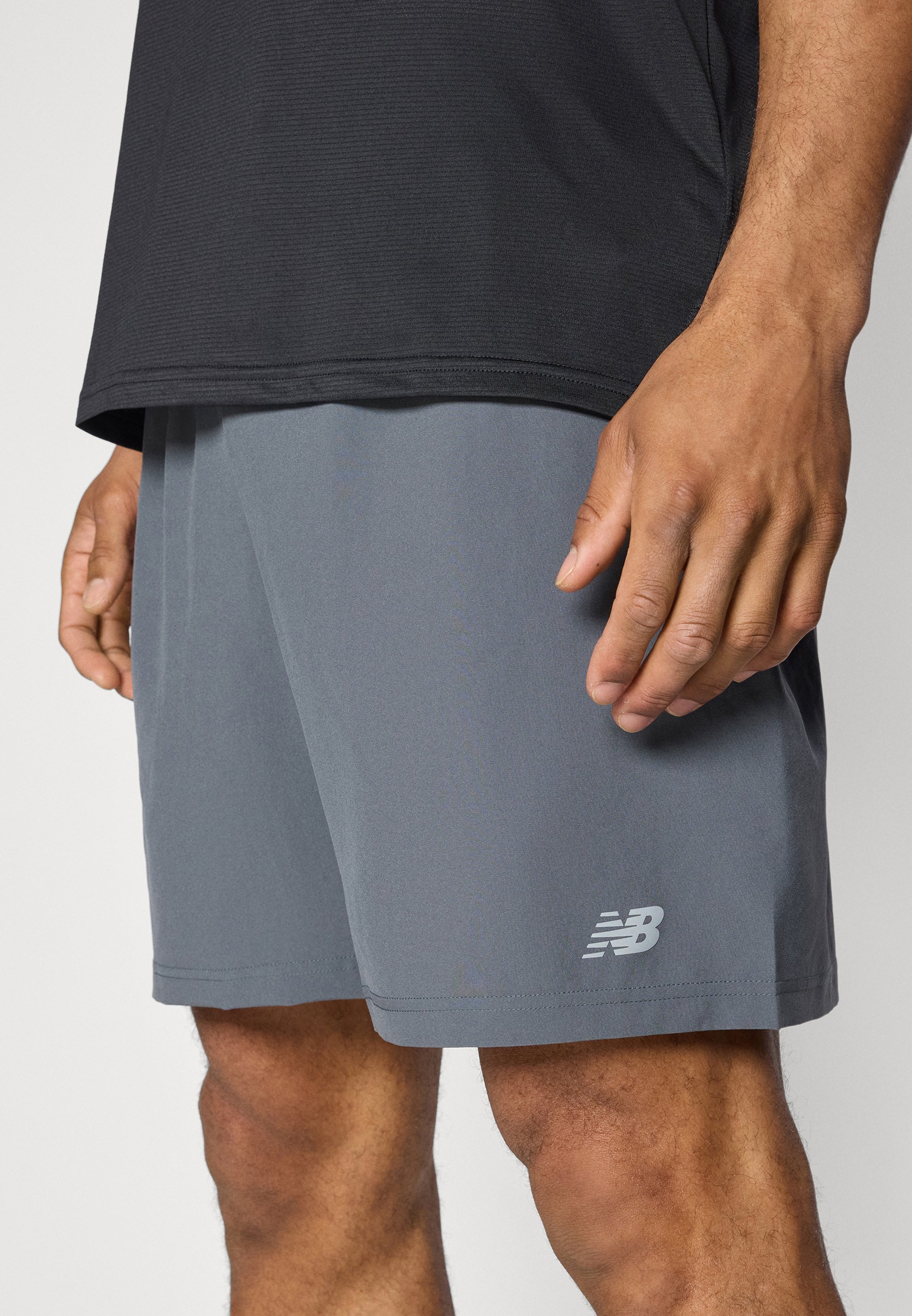 Hombre vestido con pantalones cortos deportivos grises con el logo de New Balance cerca del dobladillo y una camiseta negra, de pie con las manos relajadas a los lados.