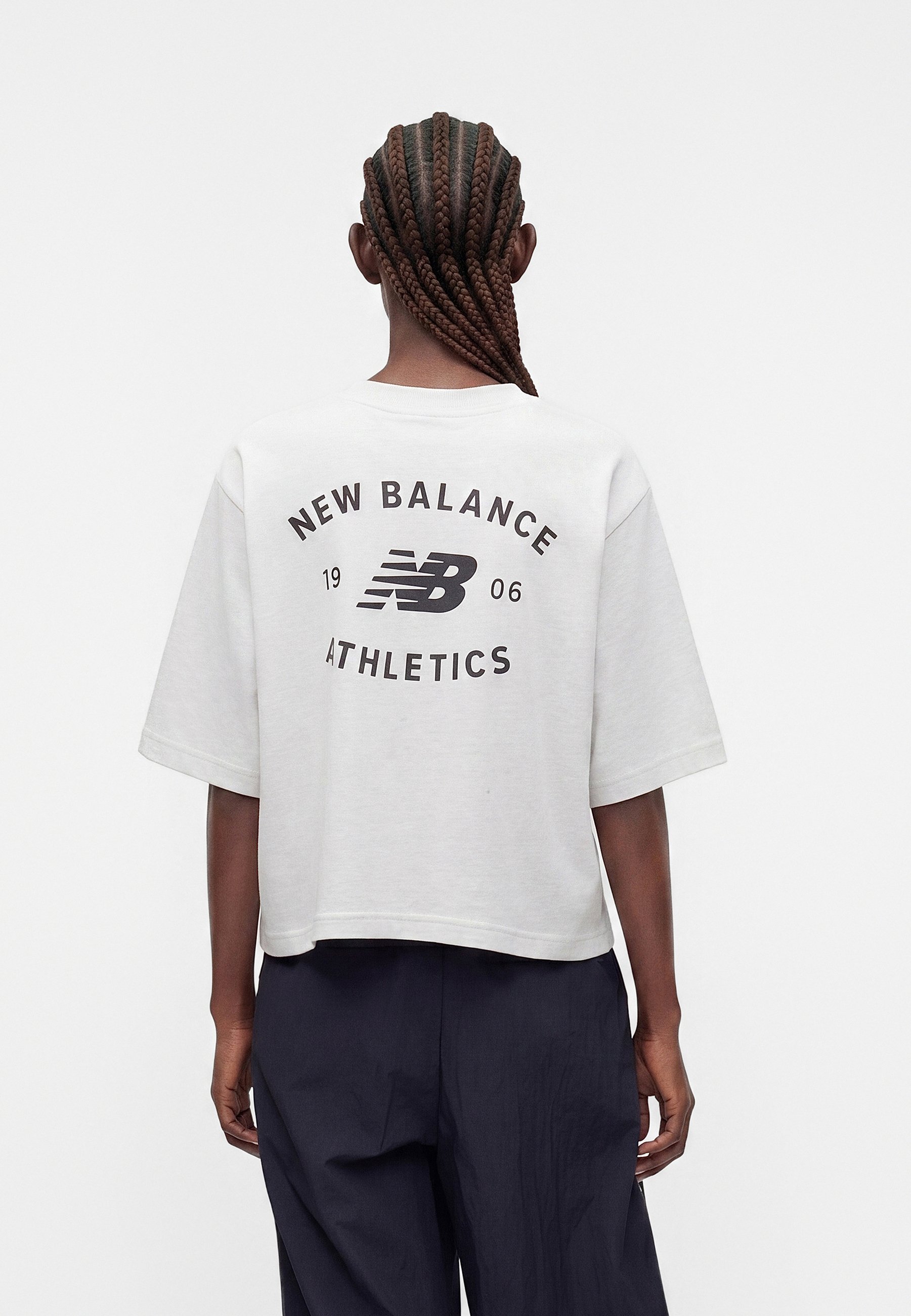 Persona con cabello trenzado que lleva una camiseta blanca corta con el logo "New Balance 1906 Athletics" en la espalda y pantalones oscuros y sueltos.