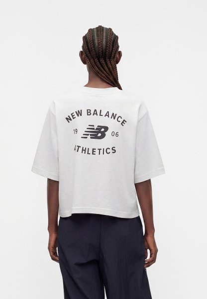Persona con cabello trenzado que lleva una camiseta blanca corta con el logo "New Balance 1906 Athletics" en la espalda y pantalones oscuros y sueltos.