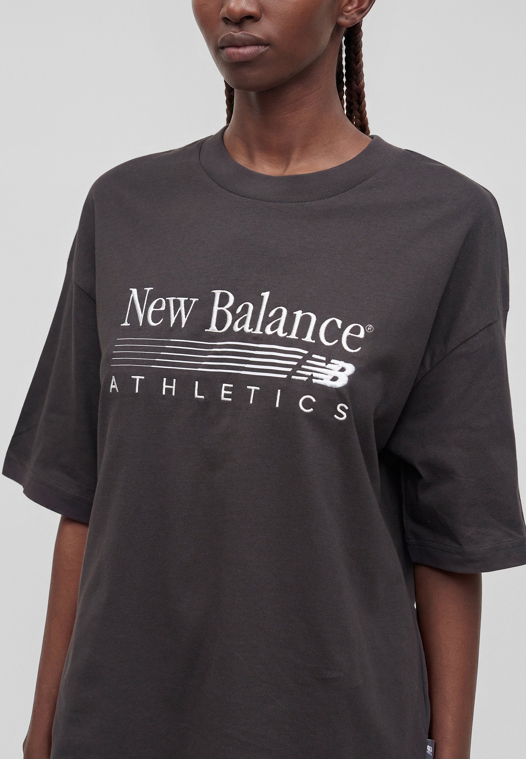 Persona con una camiseta extragrande gris oscuro con "New Balance Athletics" y logo en blanco en la parte delantera, de pie frente a un fondo liso.