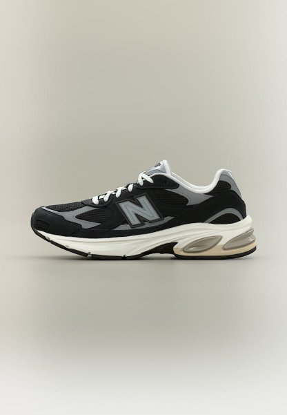 Zapatilla New Balance negra y gris con cordones blancos y suela acolchada, vista de lado sobre un fondo neutro.