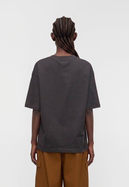 Persona vista desde atrás con el pelo largo trenzado, vistiendo una camiseta suelta de color gris oscuro y pantalones marrones holgados sobre un fondo liso.