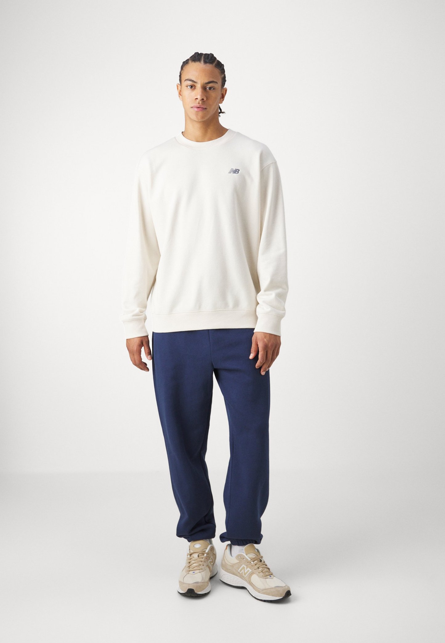 Sudadera de color crema con mangas largas, puños de canalé y un pequeño logo en el pecho, combinada con pantalones azul marino y zapatillas beige.