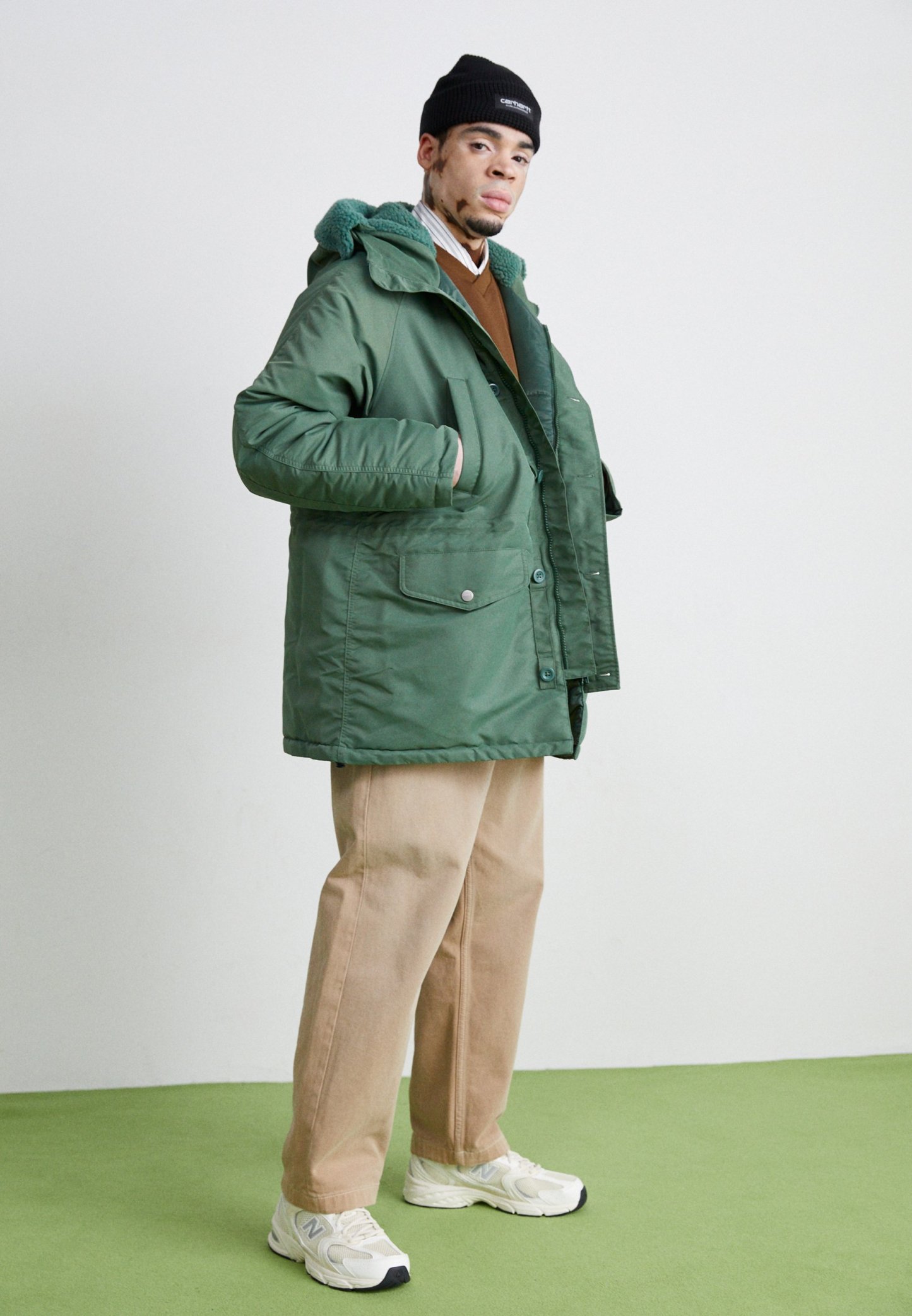 Hombre con gorro negro, chaqueta con capucha verde, suéter marrón, pantalones beige y zapatillas blancas, de pie sobre un suelo verde contra una pared clara.