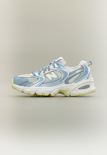 Zapatilla New Balance blanca y azul claro con paneles de malla, cordones y suela con tono amarillento sobre fondo beige liso.
