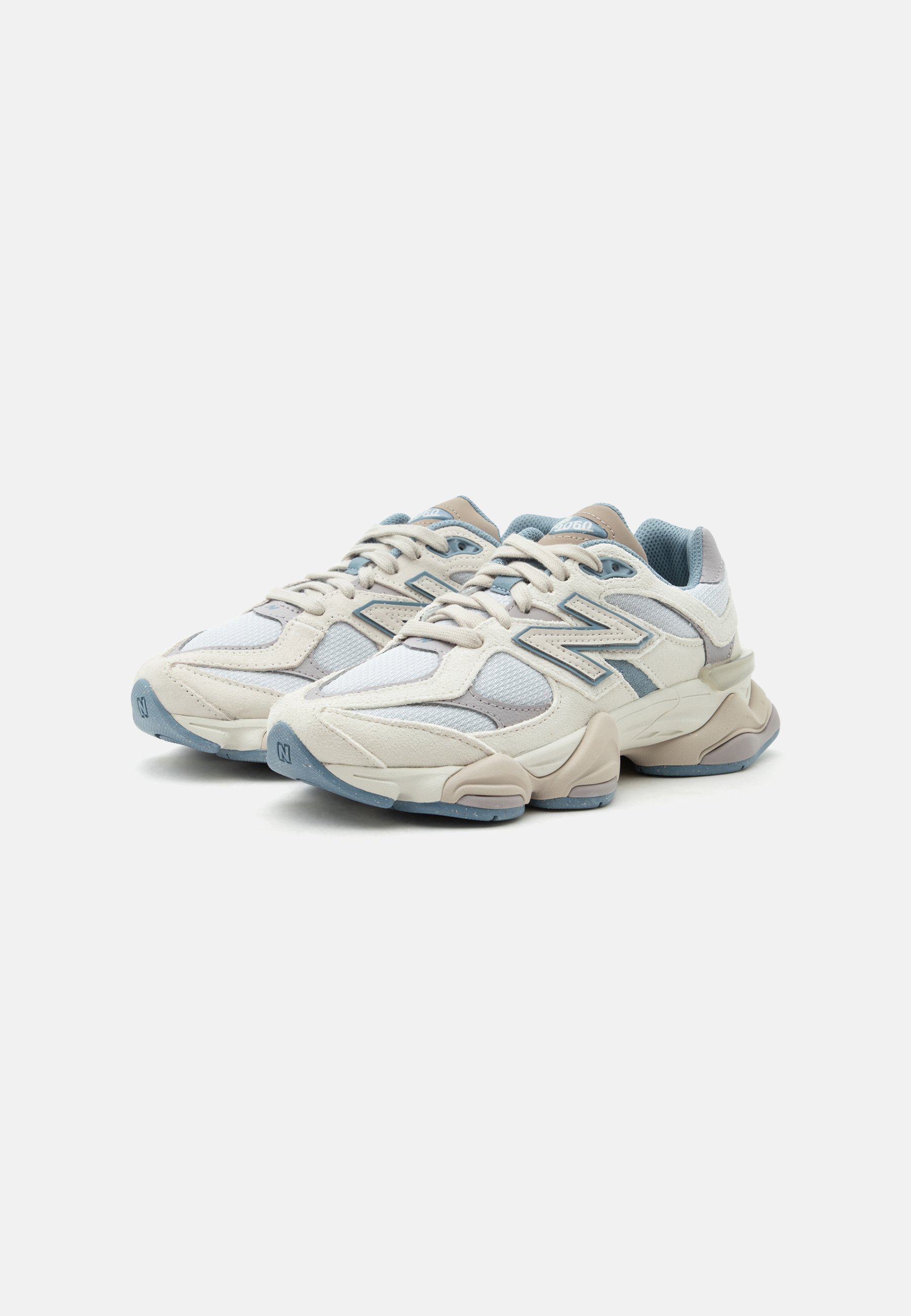 Par de zapatillas New Balance beige y azul claro con paneles de malla y ante, suela gruesa y cierre de cordones, en ángulo hacia la derecha sobre fondo liso.