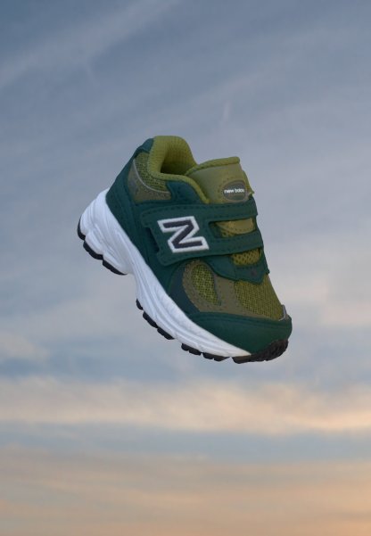 Zapatilla New Balance verde y verde azulado oscuro con suela blanca y tiras de velcro flotando sobre un fondo de cielo nublado.
