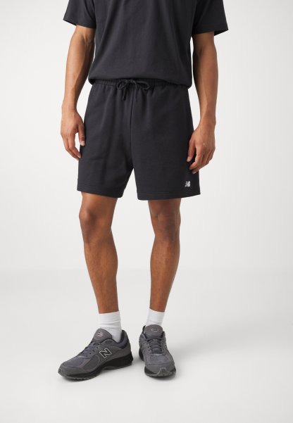 Pantalones cortos de algodón negros con cinturilla elástica y cordón, combinados con zapatillas grises que tienen un logo prominente y calcetines blancos hasta el tobillo.