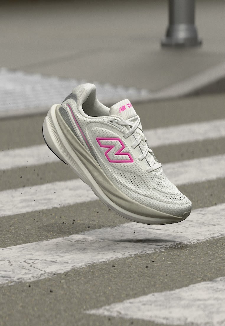 Zapatilla de running New Balance blanca y rosa en el aire sobre un paso de peatones en una carretera asfaltada.