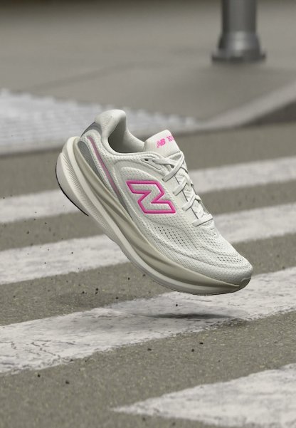 Zapatilla de running New Balance blanca y rosa en el aire sobre un paso de peatones en una carretera asfaltada.