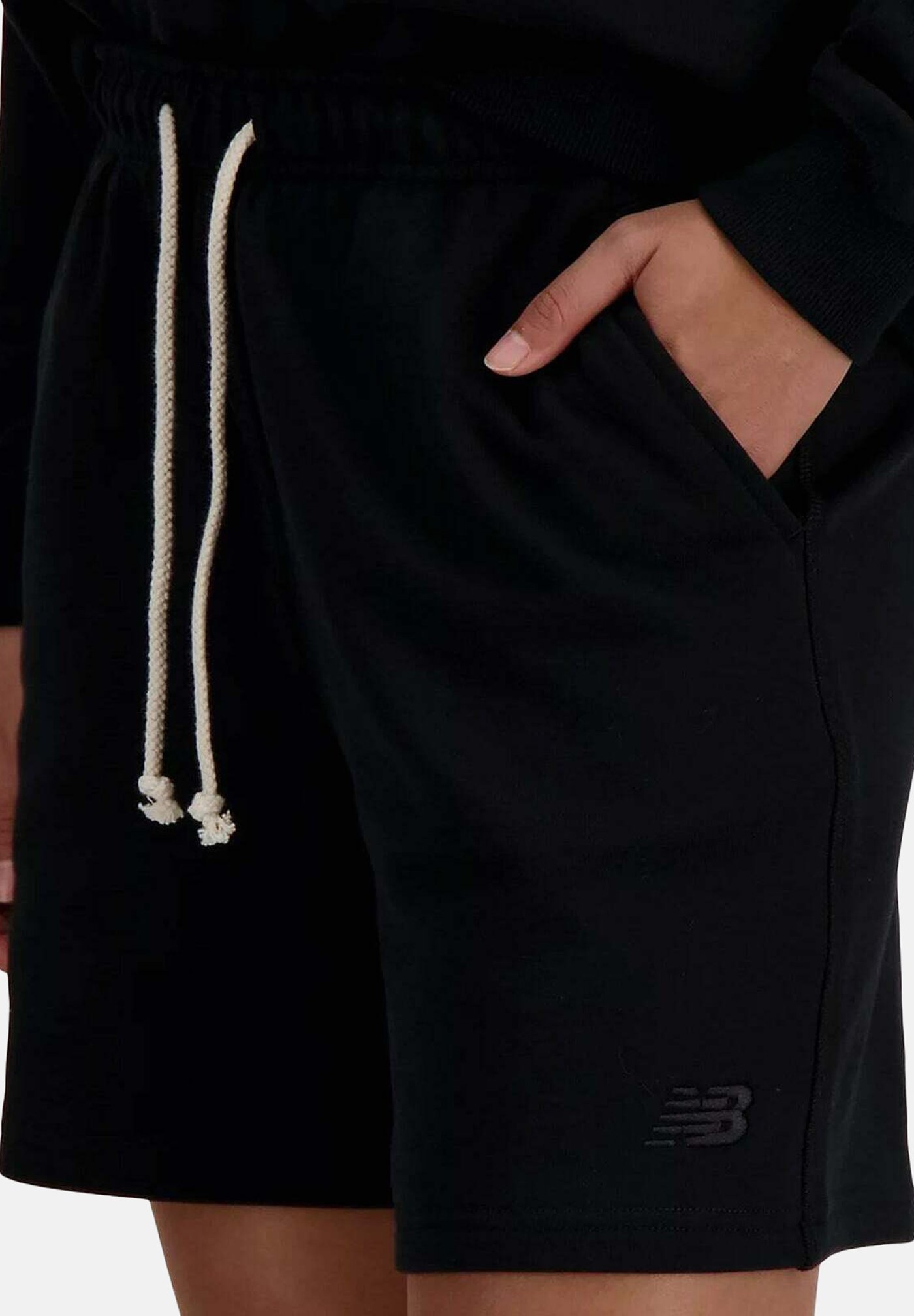 Persona que lleva pantalones cortos negros con cordones blancos, mano en el bolsillo lateral y logo de New Balance cerca del dobladillo.